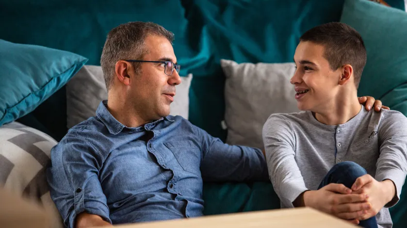 A father sits on the floor next to his teenage son, having a conversation with him.